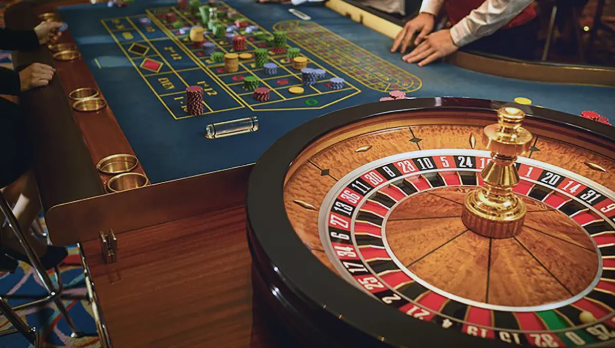 Casino roulette table scene with a close-up of the spinning roulette wheel in the foreground and the betting layout covered with chips as a dealer and players place their bets.